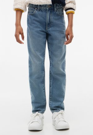 Barn iført blå denim jeans, hvide sneakers og en cremefarvet striktrøje med navy og sennepsfarvede striber på manchetterne.