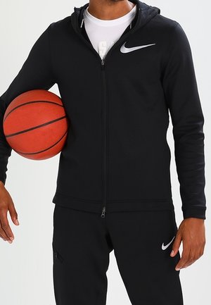 Mężczyzna w czarnym dresie Nike trzymający pomarańczową piłkę do koszykówki pod lewym ramieniem na jednolitym tle.