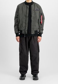 Donkergroene bomberjack met ritssluiting, geribbelde manchetten en een patch op de borst. Draagt zwart cargobroek en zwarte sneakers.