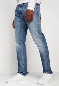 Jeans in denim blu chiaro con dettagli strappati sulle gambe, vestibilità dritta, indossati con scarpe da ginnastica bianche. Design e texture minimalisti.