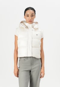 Puffervest i lys beige med hette, med kvilte seksjoner, høy krage og en trekantet logo på brystet. Glatt tekstur.