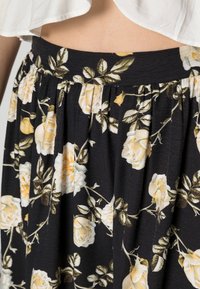 Kvinna i svart kjol med ett gult och vitt blommönster och en vit kort topp som visar en del av magen.