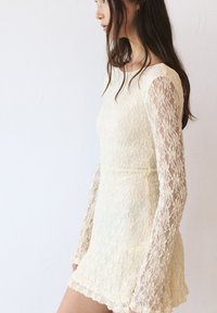 Robe en dentelle jaune pâle avec un corsage ajusté et une jupe évasée. Manches longues en dentelle transparente avec des motifs floraux et un design texturé.