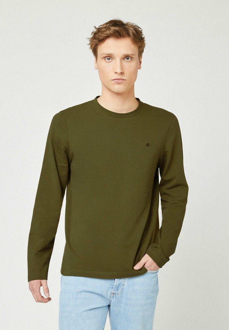 Jonge man met lichtbruin haar, gekleed in een olijfgroene lange mouwshirt en lichtblauwe jeans, staande met één hand in zijn zak.