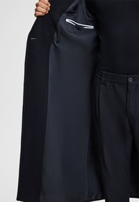 Marineblauer Mantel mit glattem, gefüttertem Innenraum, ausgestattet mit einer taschen mit Knopf und einem kontrastierenden Innenstreifen. Taillierte Form, begleitet von schwarzen Hosen.