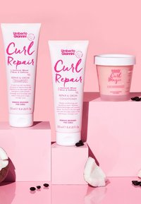 Drie roze en witte haarverzorgingsproducten op een roze display: tubes van shampoo en conditioner; een ronde container met haarmasker; kokosnootstukjes in de buurt.