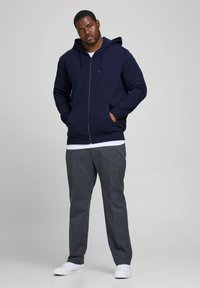 Marineblaues Zip-Hoodie mit einer Fronttasche und Kordelzugkapuze, kombiniert mit grauen gemusterten Hosen und weißen Sneakers, präsentiert auf einem neutralen Hintergrund.