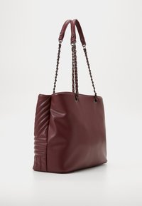 Sac fourre-tout bordeaux avec une texture lisse, doté de chaînes en tant que poignées et d’un design de panneaux latéraux matelassés pour plus de détail et de profondeur.
