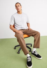 T-shirt gris à manches courtes, jean marron ample, baskets noires, chaussettes blanches, assis sur une chaise de bureau orange avec un sol vert.
