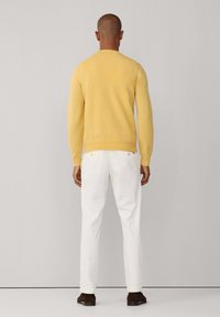 Un pull en tricot jaune à manches longues, associé à un pantalon blanc et des chaussures marron. La vue de dos met en avant la texture lisse du vêtement.