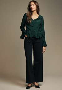 Top peplum di un verde scuro e testurizzato con scollatura a scallop e chiusura a bottoni; abbinato a pantaloni neri a vita alta e a zeppe a punta.