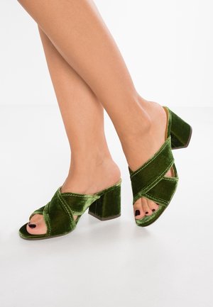 Mules de tacón de terciopelo verde con diseño de punta abierta, que presentan tiras cruzadas y un tacón grueso, mostrando una textura suave y un color rico.