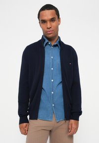 Marineblå cardigan med ribbestrikkede mansjetter, båret over en lys blå denimskjorte. Merkelig glidelåsdetalj og en liten logo på cardiganen.