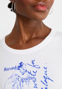 Hvid ribbet t-shirt med blå grafisk tekst og illustration, der detaljerer "Records" og "Cassettes" på forsiden. Enkel rund halsudskæring.