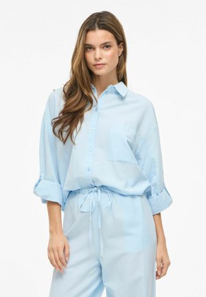 Mlada žena s dugom smeđom kosom, odjevena u labavu svijetloplavu košulju na kopčanje i odgovarajuće hlače s vezicom, stoji na bijeloj pozadini.