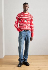 Roter Pullover mit Weihnachtsmann- und Schneeflockenmuster, Rundhalsausschnitt, gerippte Bündchen, kombiniert mit hellblauen Jeans und schwarzen Schuhen auf einem Holzfußboden.