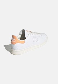 White sneaker with a smooth leather upper, orange accent on the heel, and perforated branding. Features flat laces and a textured sole.
