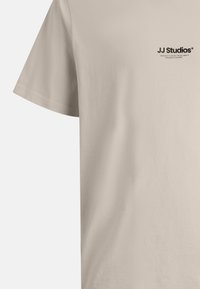 Beige Baumwoll-T-Shirt mit kurzen Ärmeln, ausgestattet mit einem schwarzen "JJ Studios"-Logo im oberen linken Brustbereich. Glatte Textur, normale Passform.