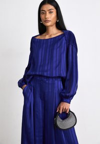 Blouse à manches longues et pantalon large en satin bleu profond avec des rayures verticales. Tenant un petit sac noir décoré de strass.