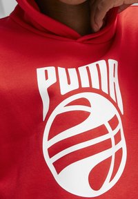 Červená bavlněná mikina s kapucí s výrazným bílým logem Puma a grafikou basketbalu, charakterizovaná hladkou texturou a atletickým designem.