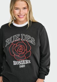 Mujer sonriente con cabello rubio que lleva una sudadera negra con una rosa roja y el texto "RUE DES ROSIERS 1989" sobre un fondo blanco.