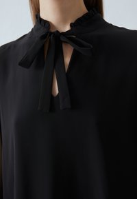 Zwarte blouse met een gerimpelde kraag, een sleutelgatopening en een strikdetail bij de halslijn. Glad stof met een zachte textuur.