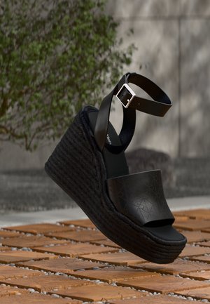 Schwarze Keilsandale mit geflochtener Plateau-Sohle und Knöchelriemen mit silberner Schnalle, auf Holz- und Kiesboden im Freien platziert.