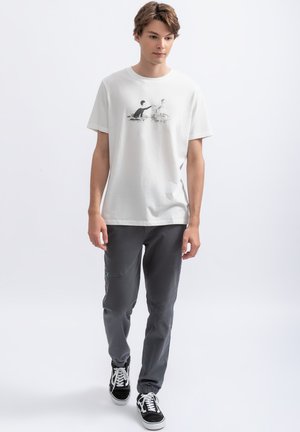 T-shirt bianca in cotone con design grafico nero raffigurante due figure. Pantaloni grigi con cuciture a contrasto e sneaker nere con lacci bianchi.