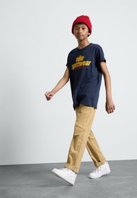 Zilas jūras zilas krāsas īsām piedurknēm T-krekls ar dzeltenu "Nike Sportswear" tekstu, apvienots ar khaki biksēm un baltām kedām, papildināts ar sarkanu beanie. 