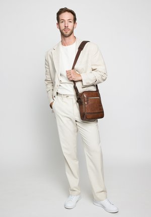 Man wearing cream suit, white shirt, white sneakers, and carrying brown leather crossbody bag.