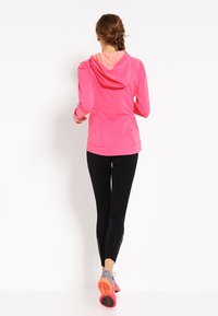 Světlá růžová sportovní bunda s kapucí a kontrastním vnitřním podšíváním, kombinovaná s černými legínami s síťovanými panely a růžovými sportovními botami.