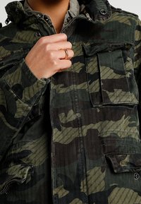 Giacca mimetica nei toni del verde scuro e nero, con chiusura a zip e bottoni automatici, diverse tasche frontali e tessuto strutturato.
