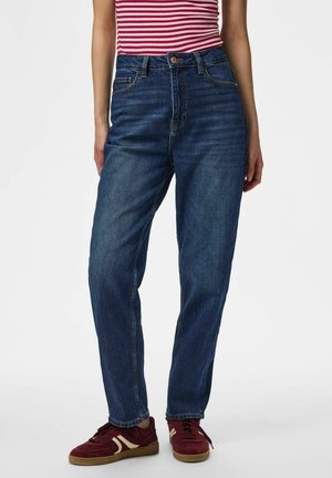 Persoon draagt blauwe jeans met een middenhoge taille, een rood-wit gestreept topje en rode sneakers, staand tegen een effen witte achtergrond.