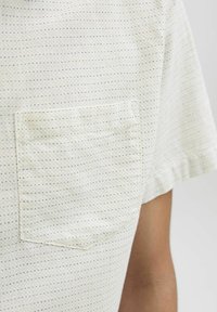 Chemise à manches courtes en tissu beige clair avec de petits pois bleus et noirs, dotée d'une poche plaquée sur la poitrine gauche.