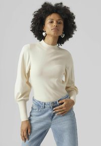 Top in maglia costina di colore crema con colletto alto e maniche lunghe a sbuffo, abbinato a jeans a vita alta di colore blu chiaro.