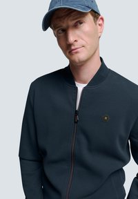 Dunkelblaues Zip-Jackett mit strukturierter Oberfläche, am Kragen geöffnet. Verfügt über ein hexagonales Logo auf der linken Brust und ein weißes Unterhemd.