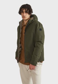 Giacca verde con cappuccio e chiusura a zip, dotata di una fodera interna trapuntata e tasche laterali. Indossata sopra una camicia marrone. Texture liscia.