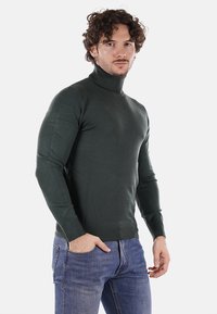 Maglione dolcevita verde scuro in un design aderente, realizzato in un materiale a maglia liscia con maniche lunghe e orlo a coste. Indossato sopra dei jeans blu.