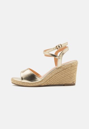 Gold metallic wedge sandal with a woven jute sole, open toe, and adjustable ankle strap. Smooth texture with a seamless design.