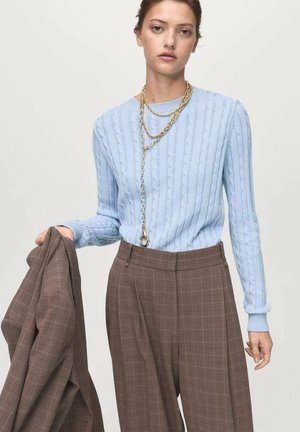 Lichtblauwe kabelgebreide trui, oversized pasvorm. Bruin geruite getailleerde broek met een hoge taille. Gouden kettinghalsspeld rondom de nek.
