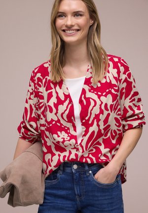 Femme souriante aux cheveux blonds portant une chemise rouge et beige à motifs sur un haut blanc, jean bleu, tenant une veste beige pliée.