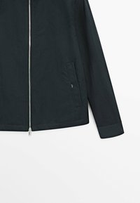 Giacca nera con maniche lunghe, cerniera frontale argentata e tasca laterale con bottone a pressione, mostrata su sfondo bianco.