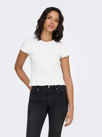 Bijela rebrasta kratka majica s valovitim rubovima, nošena s crnim trapericama. Tkanina ima teksturiranu završnu obradu, stvarajući prijanjajuću siluetu.