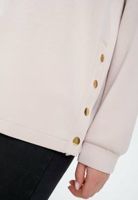 Lichtroze sweatshirt met een zachte textuur, voorzien van gouden knopen aan de zijkant en een lichte zijsplit aan de zoom. Zwarte broek zichtbaar.