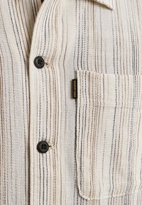 Chemise boutonnée tissée en crème avec des rayures verticales marron et noires. Comprend une poche poitrine et une étiquette de logo noire. Tissu texturé.