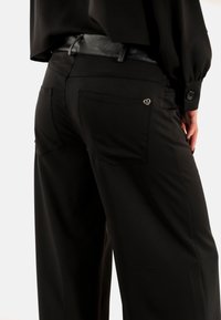 Pantaloni neri a gamba larga con una texture liscia, dotati di due tasche posteriori e un piccolo logo metallico accentato sulla tasca destra.