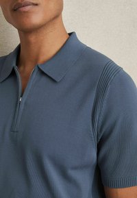 Polo cu mânecă scurtă, de culoare albastru închis, cu un aspect texturat din materiale ribbed, guler cu fermoar și detalii cu dungi verticale pe umeri.