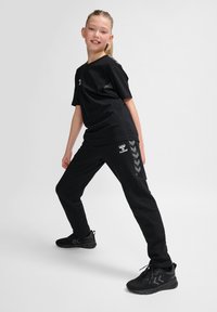 Schwarzes Baumwoll-T-Shirt und passende Jogginghose mit grauen Chevron-Akzenten. Enthält ein Markenlogo auf T-Shirt und Hose. Schuhwerk: schwarze Sportschuhe.