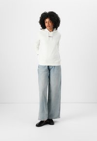Witte hoodie met een trekkoord en bedrukt logo, gecombineerd met lichtblauwe wijdere jeans en zwarte instappers. Soepele stofstructuur.