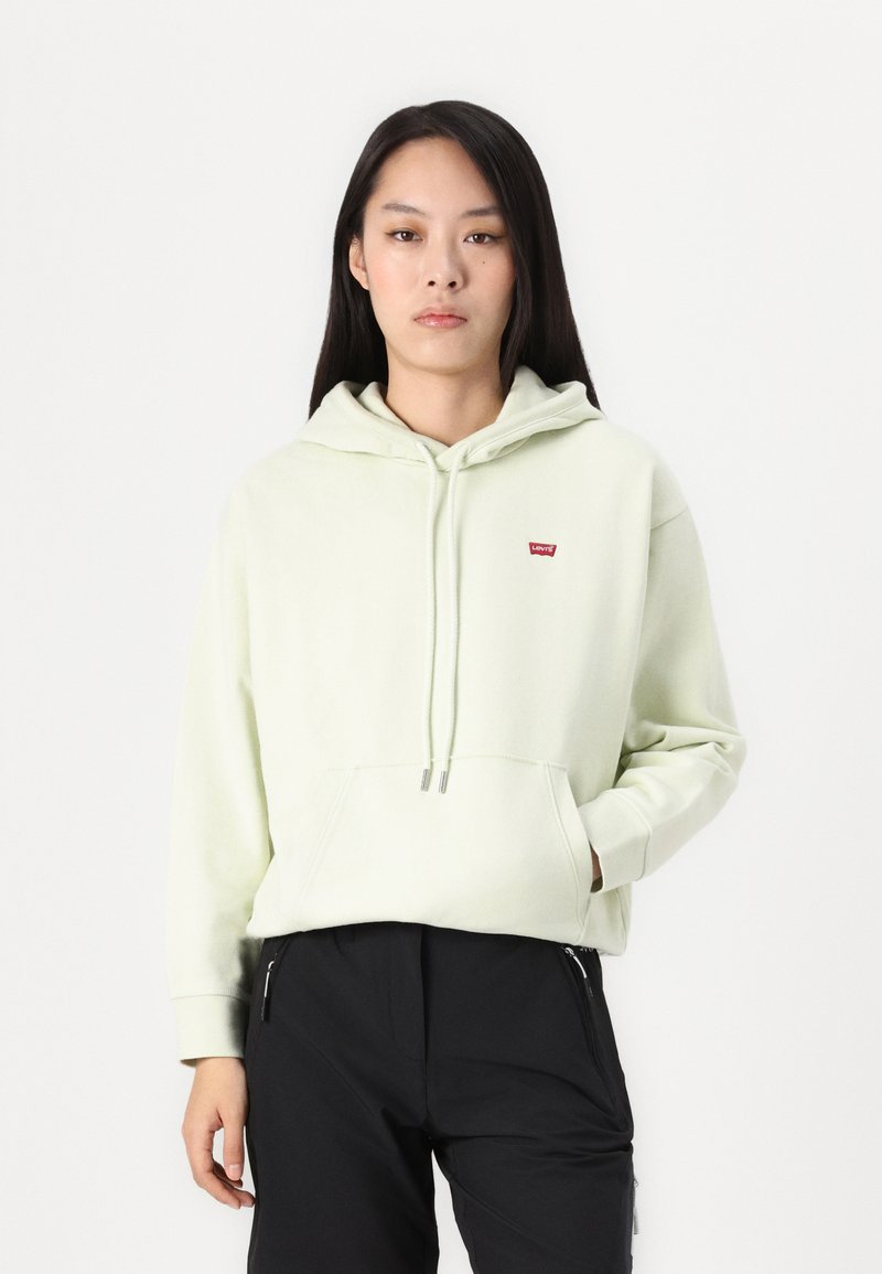 Donna che indossa un hoodie Levi's verde chiaro e pantaloni neri, in piedi davanti a uno sfondo bianco semplice con una mano in tasca.